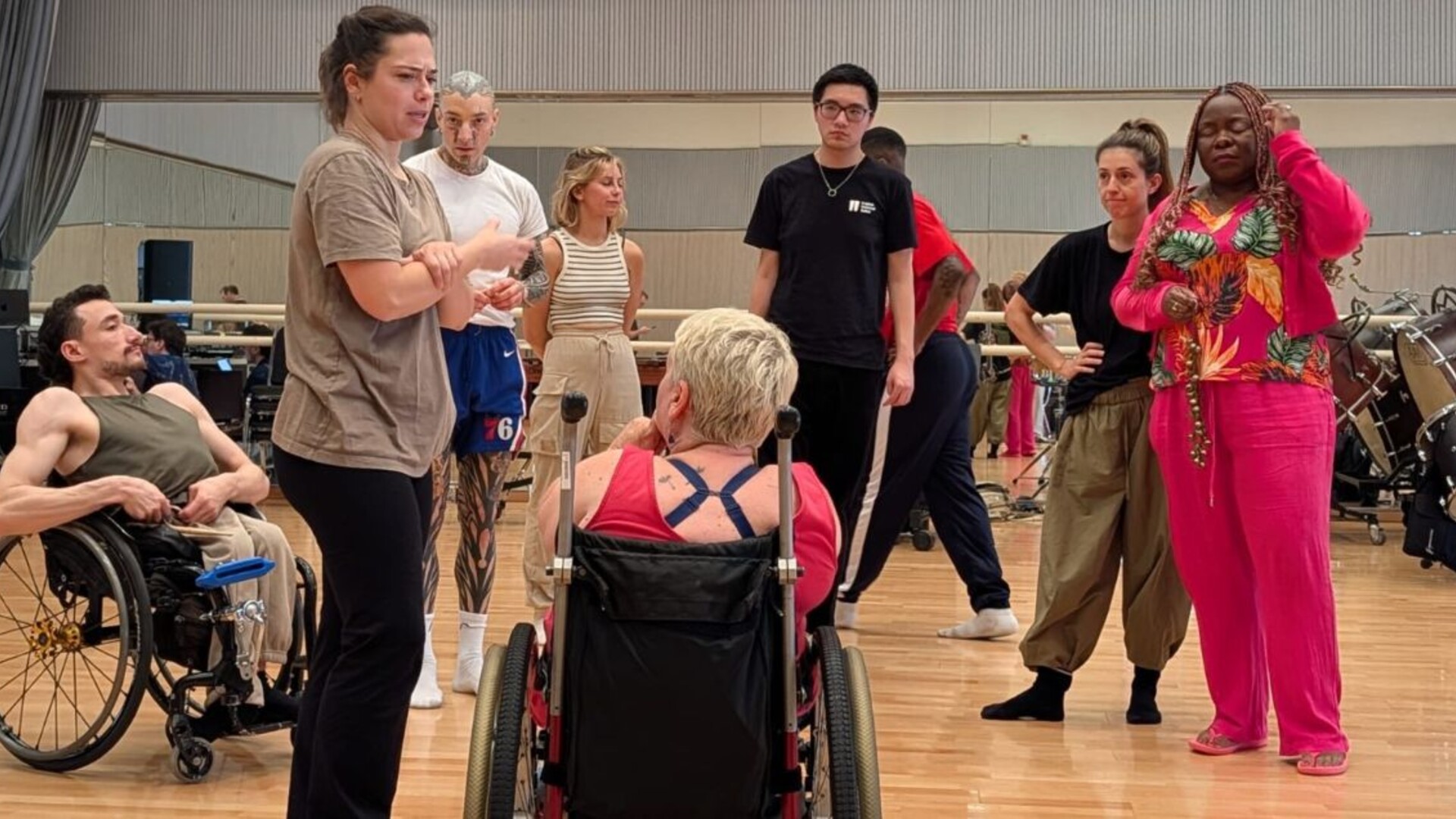 A group of dancers gathered in a rehearsal studio, standing in a circle and listening to a woman speaking.