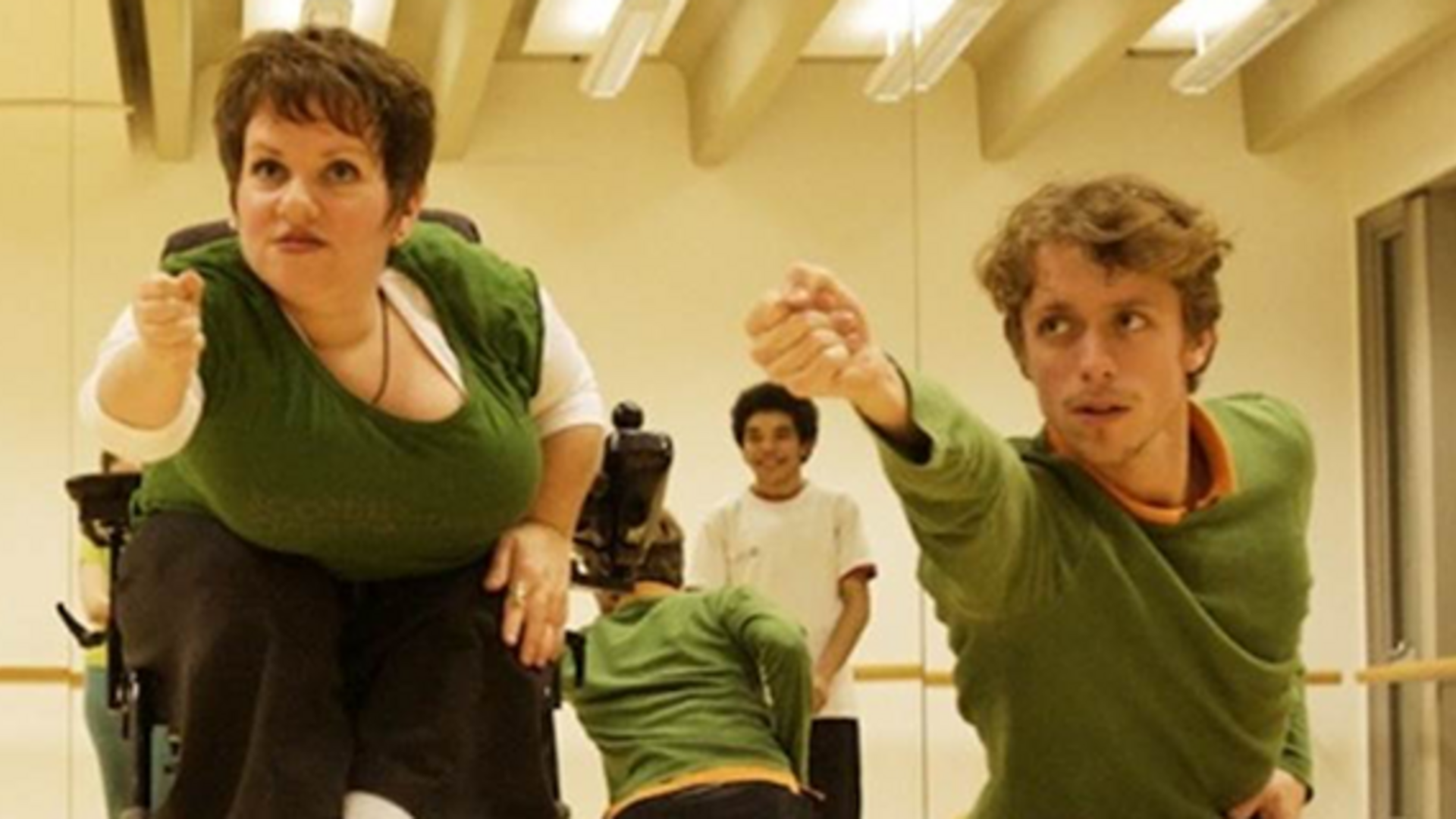 Caroline Bowditch with arm outstretched in dance studio as a person next to her holds the same pose, and a group of dancers look on