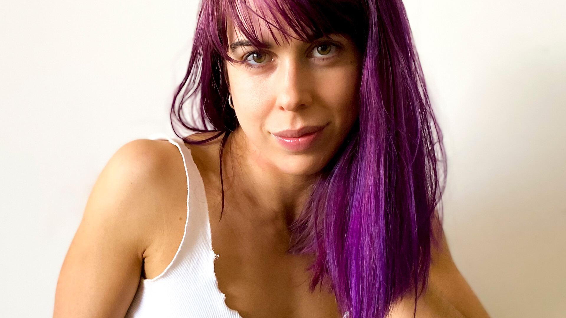 Amy is in front of a white background. She is wearing a white singlet and her head is resting on her left hand. Amy has purple hair and is looking directly at us.