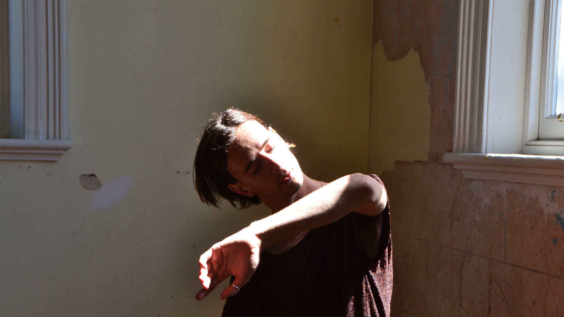 Ben is dancing in the corner of a room, the walls behind him are peeling paint. His left arm is raised, bent at the elbow and crossing in front of his body—with his palm facing away from him. His head is tilted towards the left catching the natural light coming through the window. He is wearing a brown t-shirt and a silver chain around his neck and a ring on his thumb and first finger.