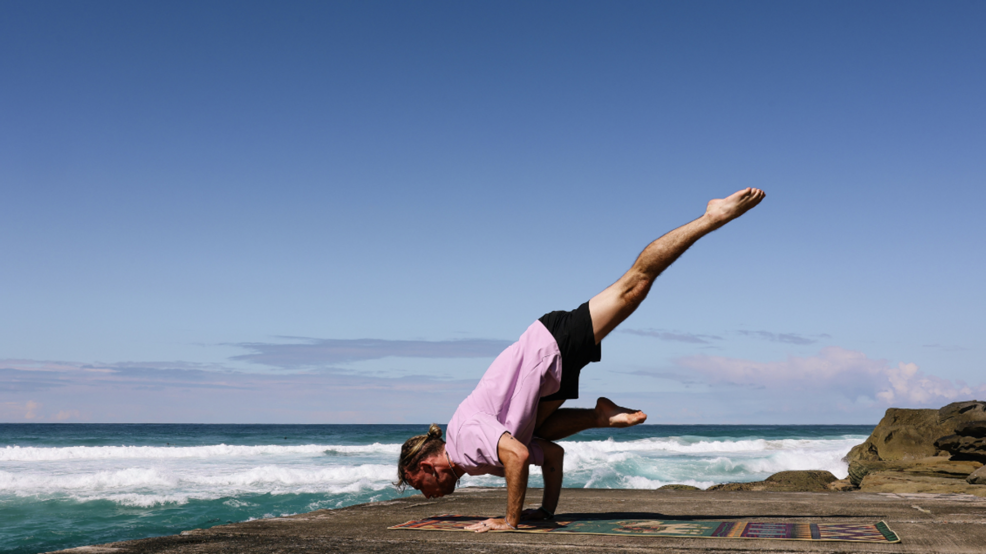 Callum is doing an arm balance on his colour yoga mat at the beach.