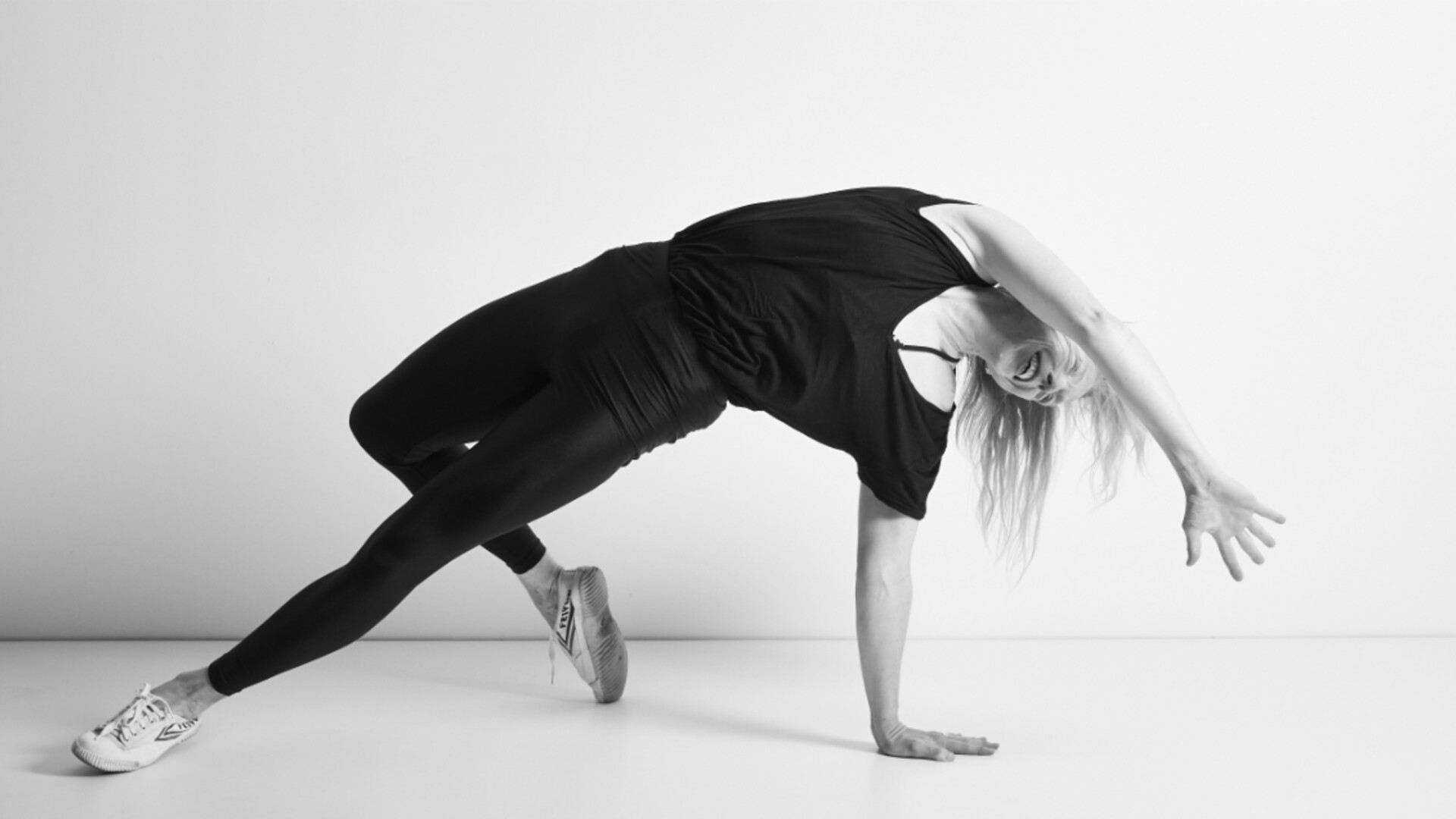 A black and white image of Jo transitioning from a yoga pose, her body is stretched out long weight supported by left arm and leg, her right leg is bent behind her right tips of toes on the floor while her right are is suspended reaching past her face away from this. She is wearing a loose black t-shirt and black tights with white lace up sneakers, she is smiling.