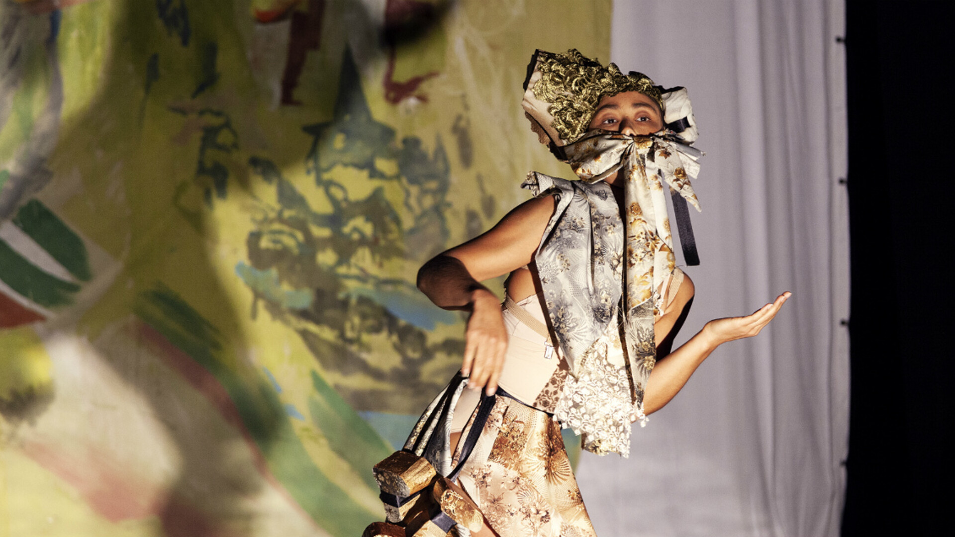 Lilian is dancing in front of a coloured backdrop painted with earth tones. Her head is tilted slightly up and towards the right while her arms are waist height bent at the elbows, left palm upwards facing towards her head, right hand limp at the wrist facing down. She is wearing a headpiece tied on in a bow across her cheeks and mouth, her torso covered with material pieces draped in layers over a light coloured tight top.