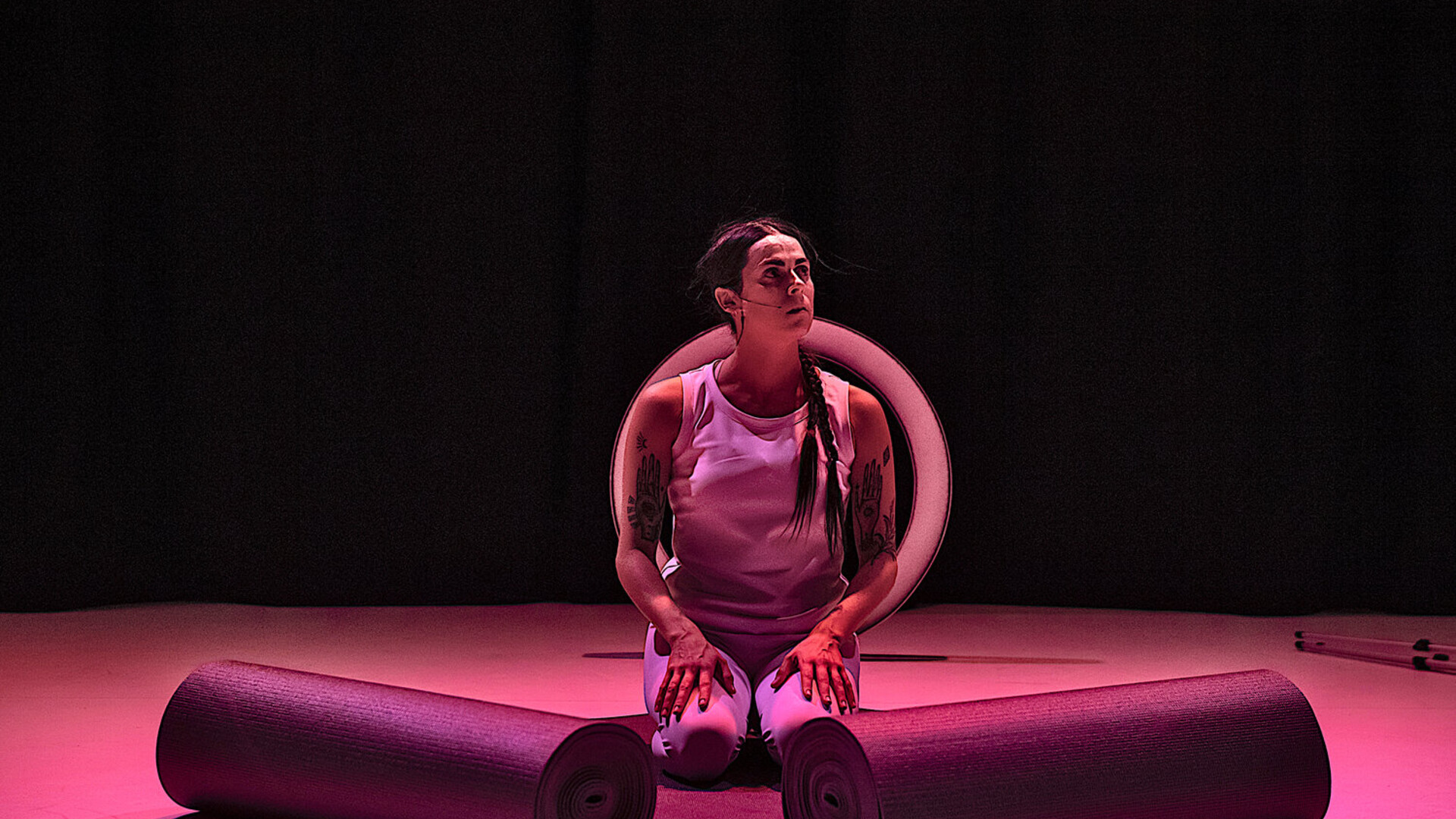 Rachel kneels on the unrolled portions of two yoga mats that lie one on top of the other. Rachel’s torso faces us, but their face is tilted away slightly upward to the right. The floor is lit in pink contrasting from the dark background.