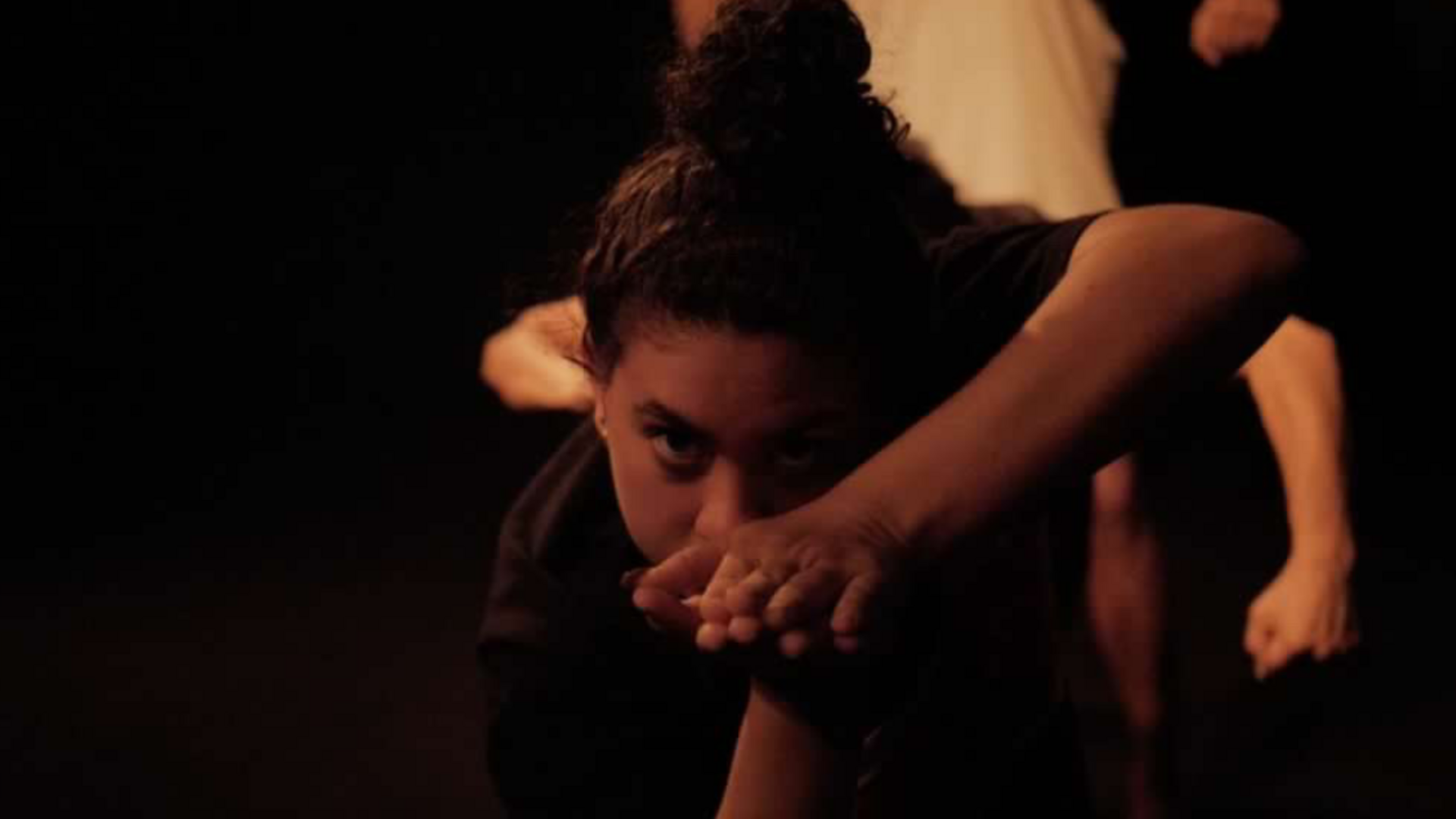 Dancer Brianna Kemmerling is performing on a dark stage. Her hands clasped together in front of her face, looking directly at you.