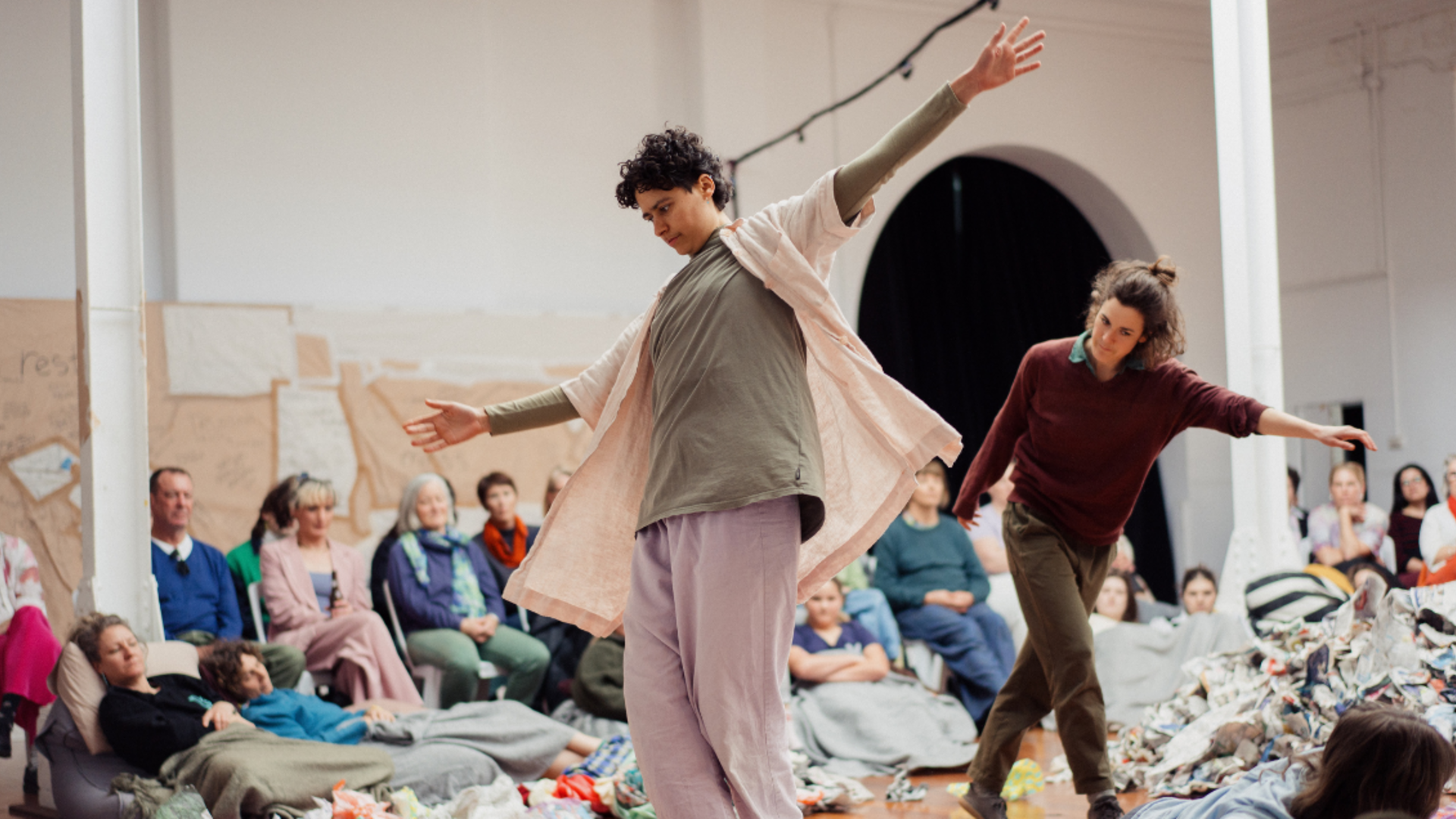 Contemporary dancers perform amid colorful scattered materials in a gallery, while seated spectators watch; the scene feels lively and expressive.
