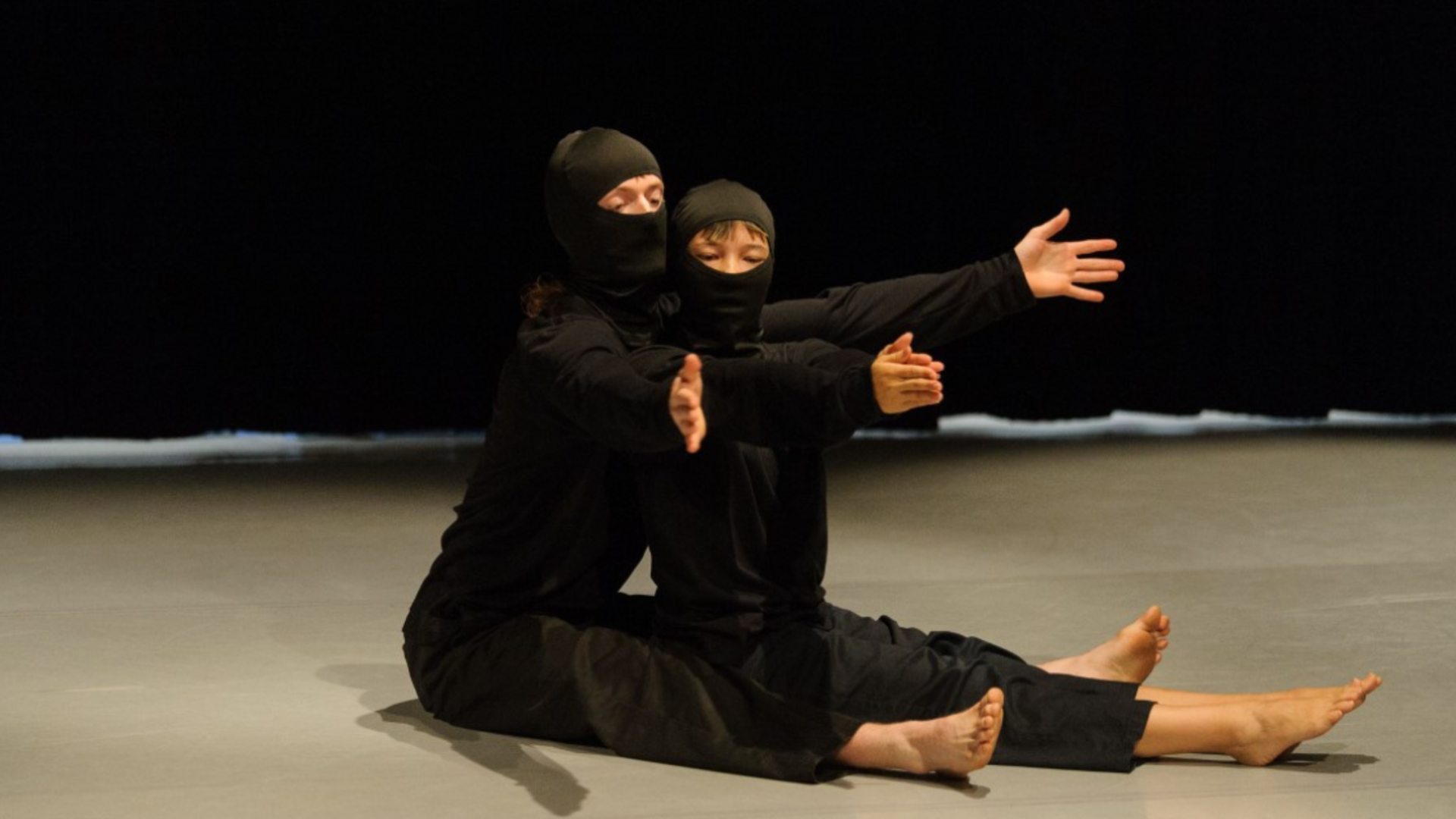 2 dancers dressed in black and wearing balaclavas sit on the floor of a dane studio, one behind the other. Both with outstretched arms and legs.