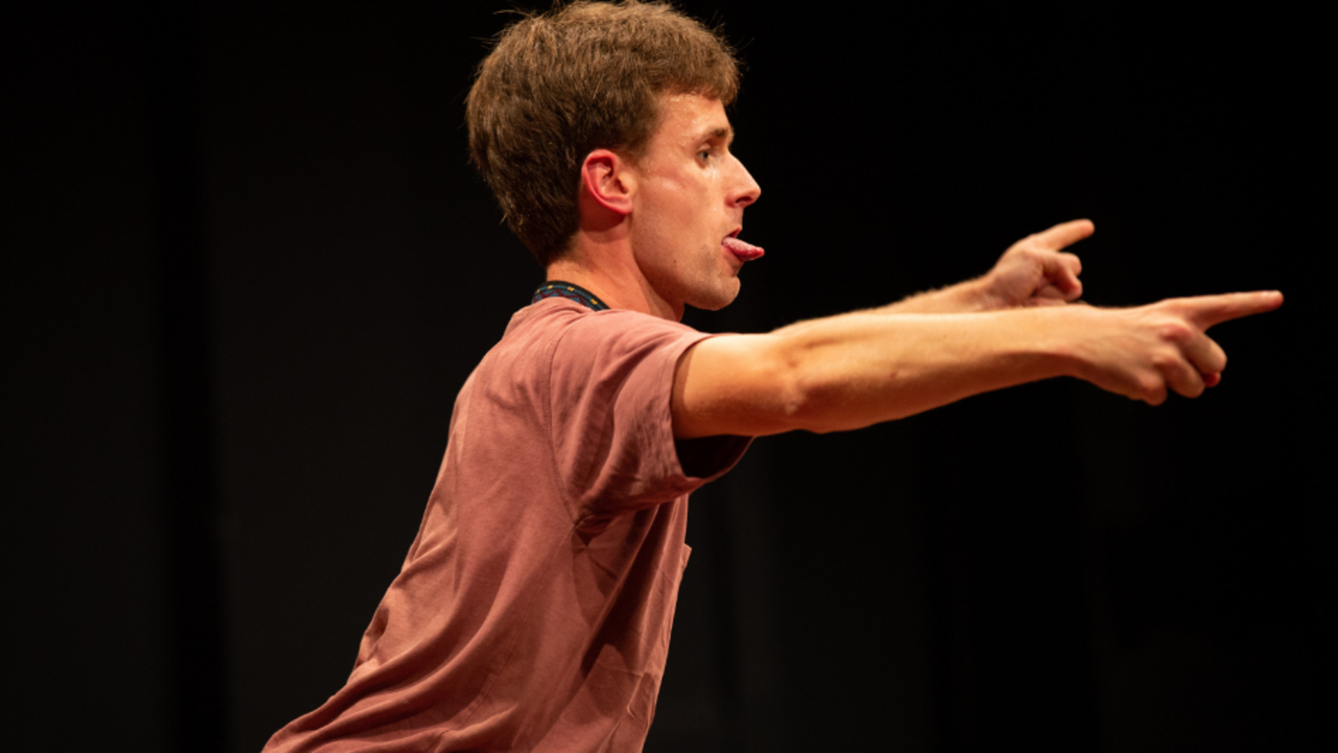 Dancer Thomas Woodman facing sideways in a dance studio. He wears a brown tshirt. He reaching his arms forwards with his fingers pointing. He is also sticking his tongue out.