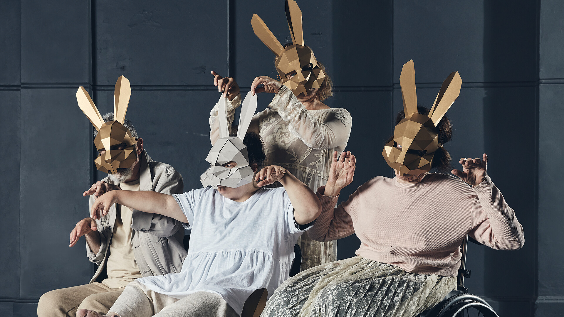 Four performers wearing geometric rabbit masks are facing toward us, arms and hands raised.