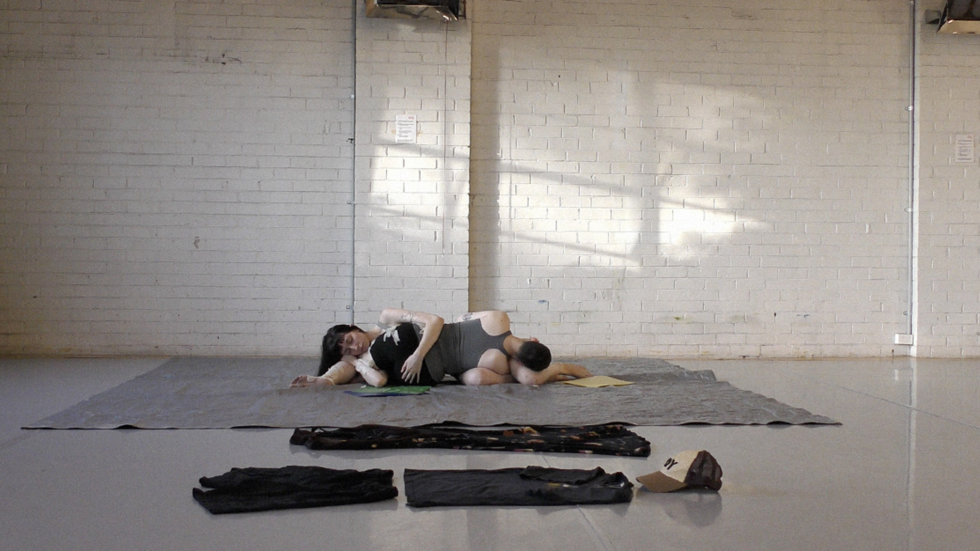 Two dancers are lying on their sides, bodies entwined, on a grey mat in a brightly lit studio.