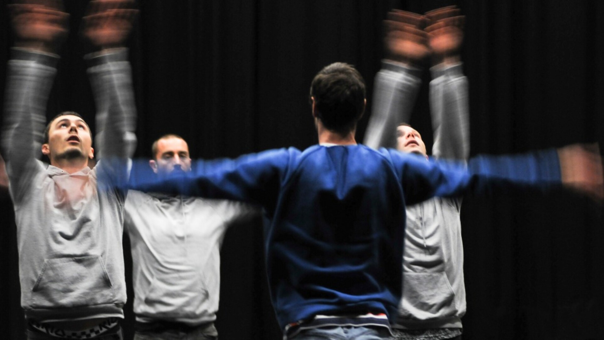 4 dancers, 3 in grey tops facing 1 in a blue top, wave their arms energetically while looking up or forward in front of a black wall
