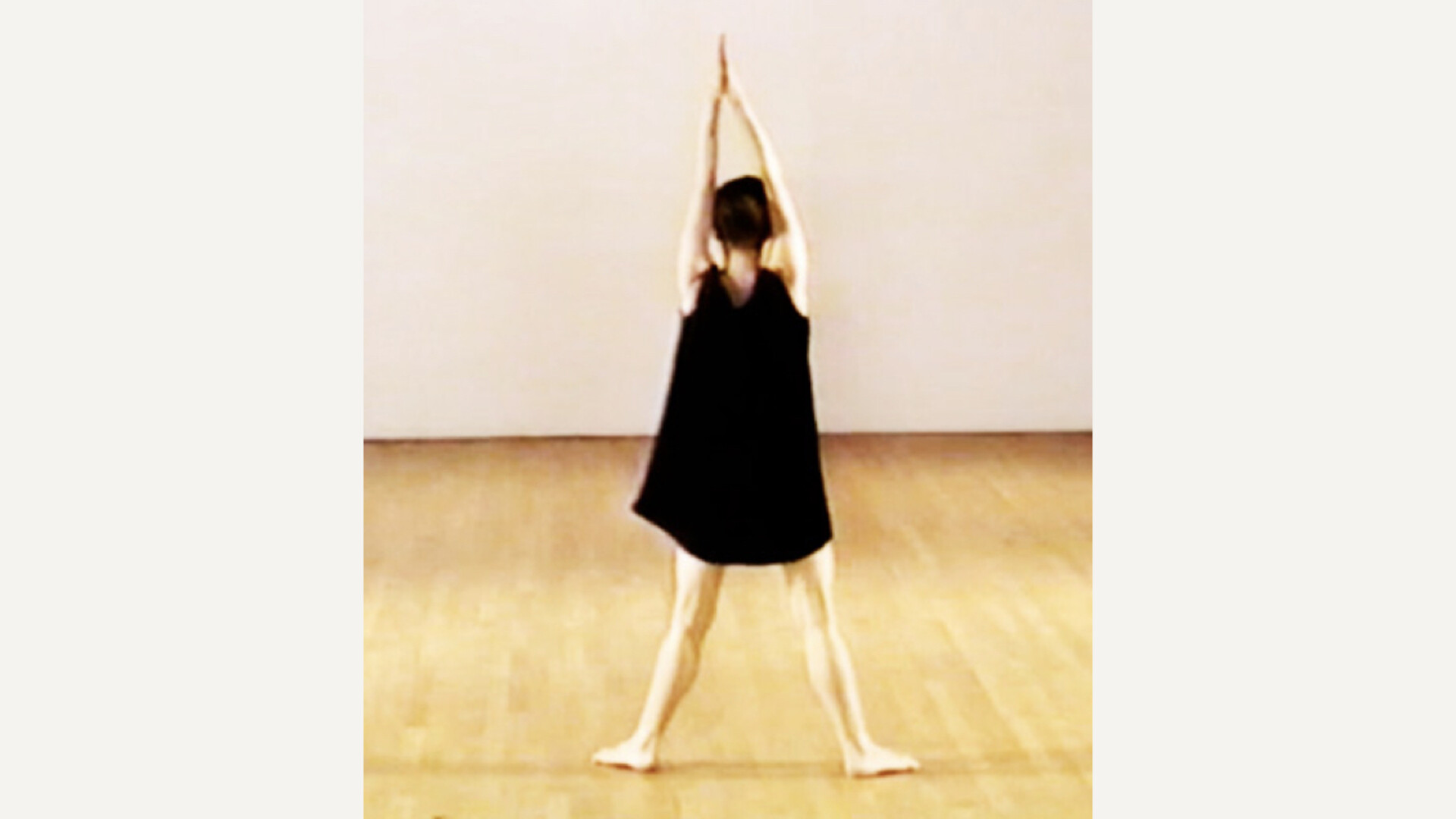 A dancer in a short black dress faces away with feet apart and arms held aloft, palms together in a large room with light wood floors and white floor