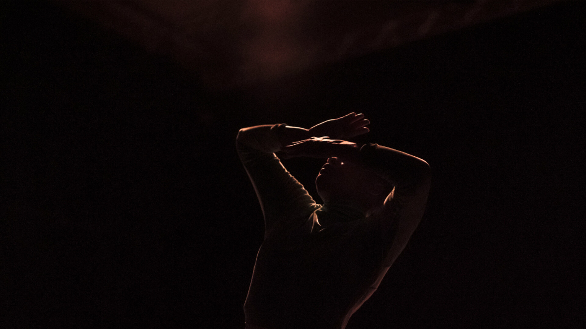 A dancer is dimly lit, holding her hands folded above her head.