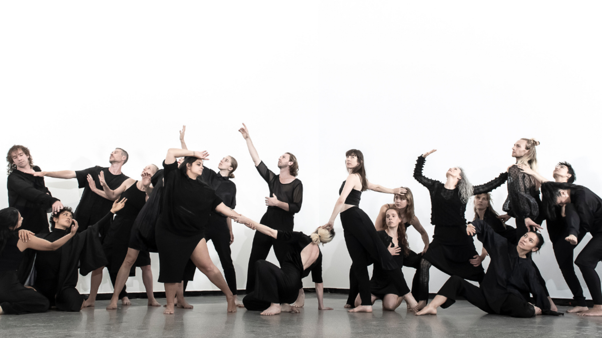 19 dancers dressed in black pose in a tableau, in a white studio with a polished concrete floor.