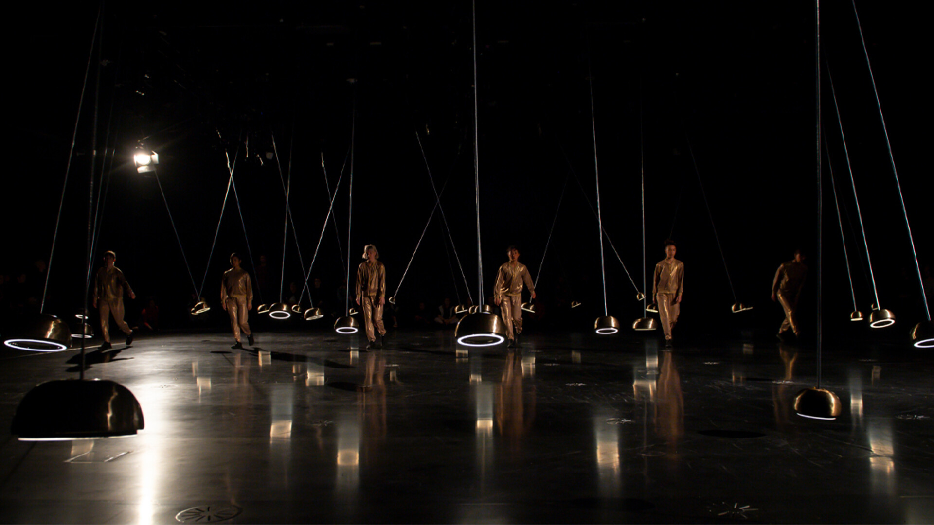 Dancers wearing gold tracksuits move in formation across a dark stage between pendulums.