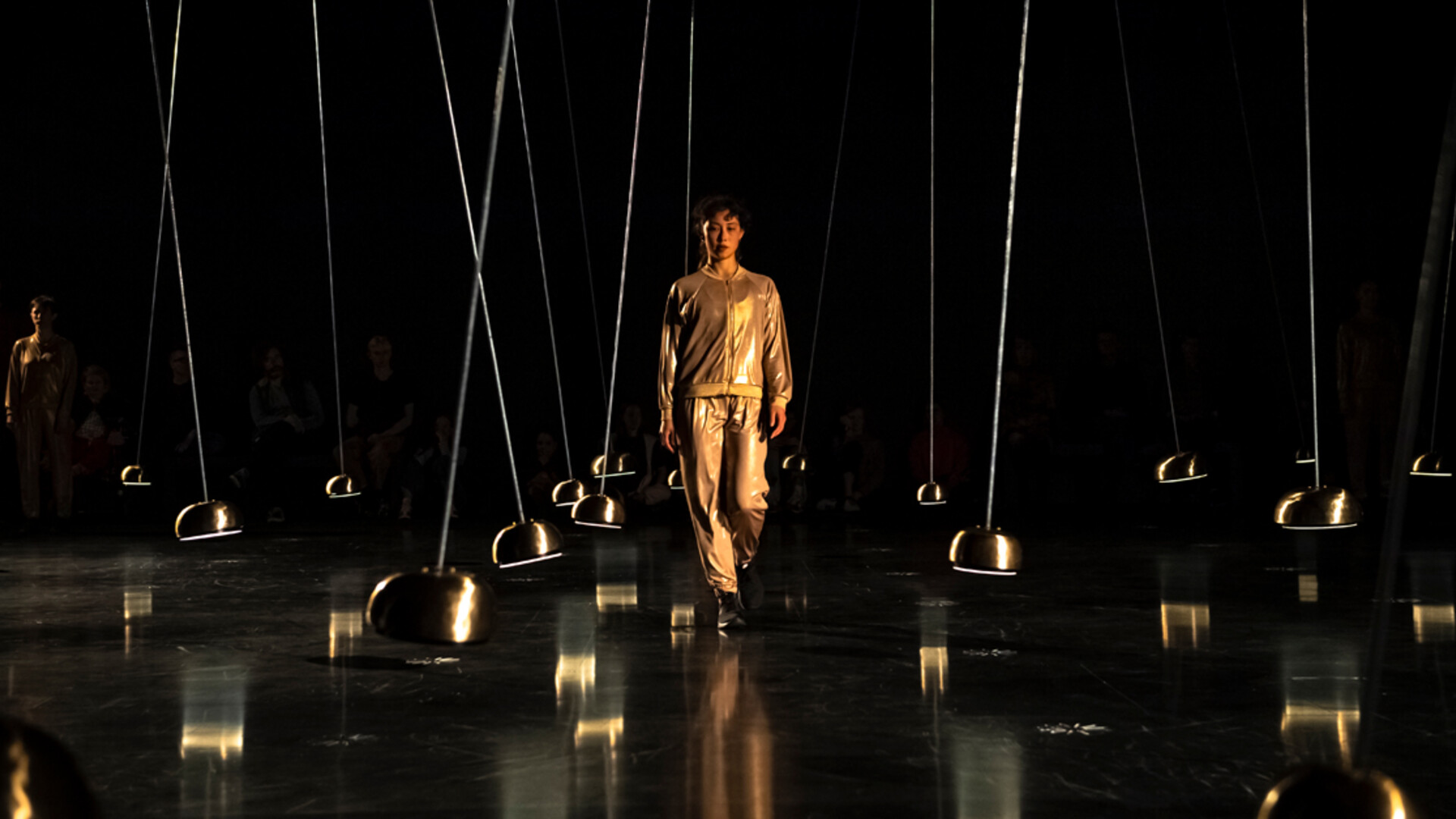 A dancer wearing a gold tracksuit walks toward the camera between suspended pendulums.