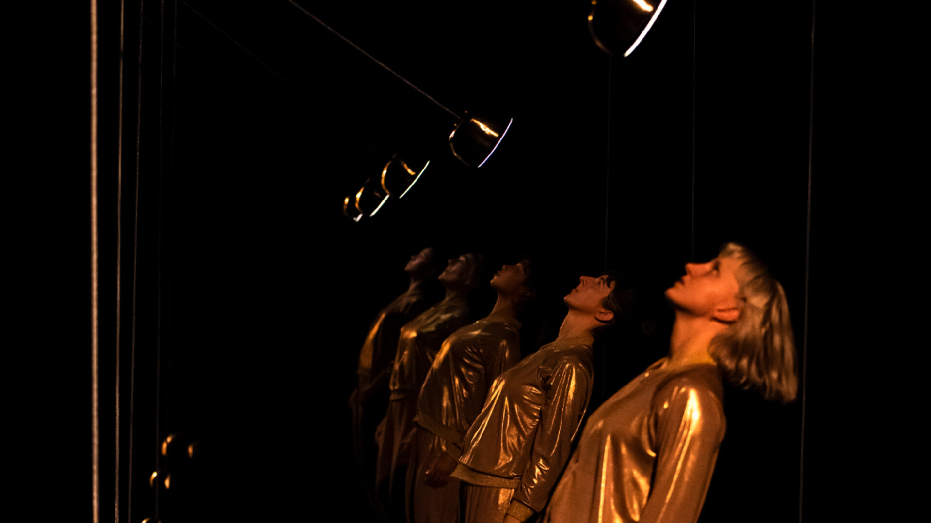 Dancers in gold tracksuits stand in a row with their heads tilted back as pendulum swing above their faces.