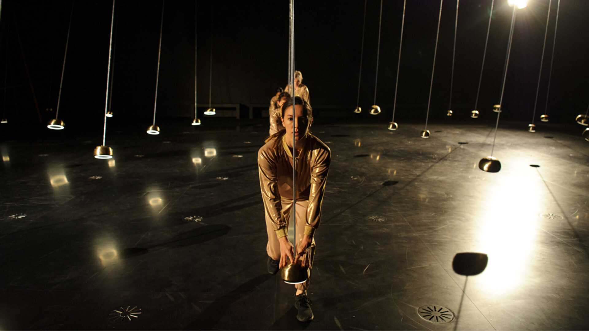 Dancers in gold tracksuits crouch in a row holding pendulums.