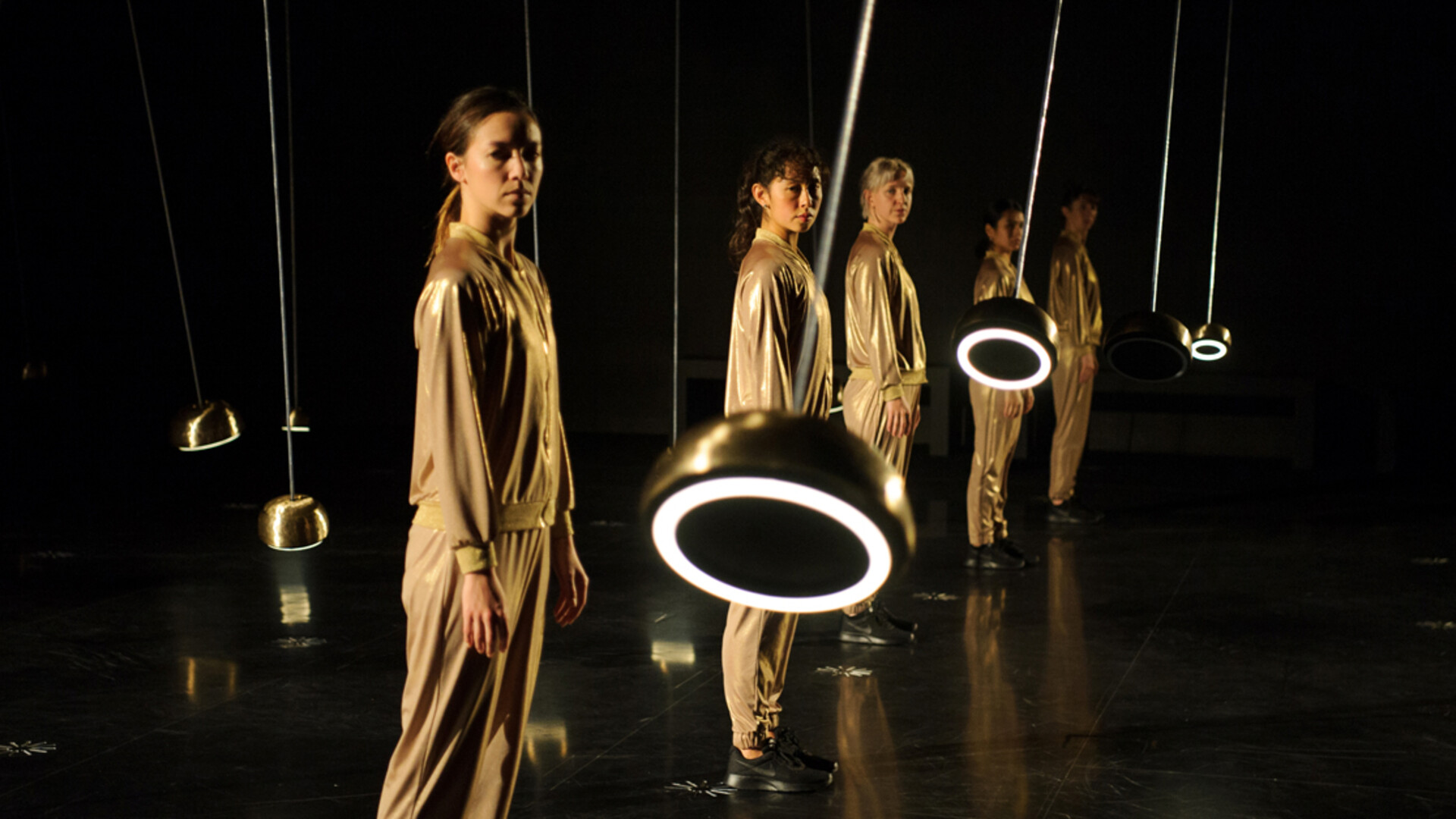 Dancers in gold tracksuits watch pendulums as they sway away from their bodies.