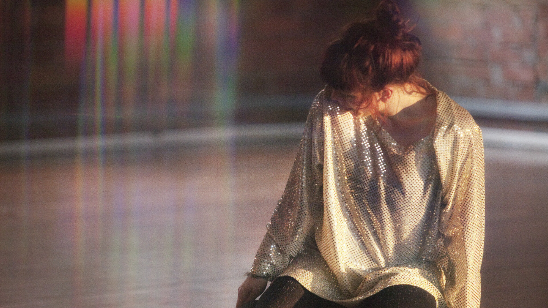 A woman kneels and looks away from the camera to her right in a sparkly dress. The light in the image creates a rainbow effect on the left