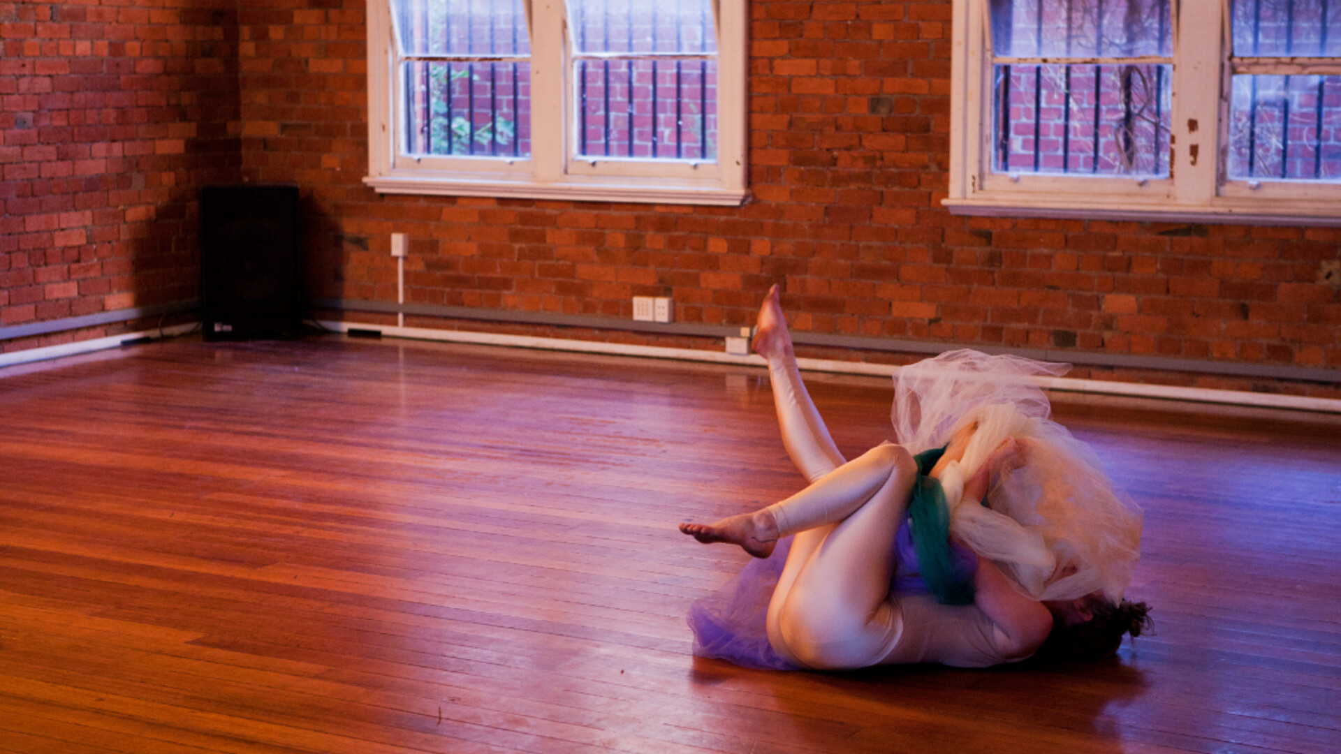 A woman lies on a wooden floor in white tights clutching material that covers her front.