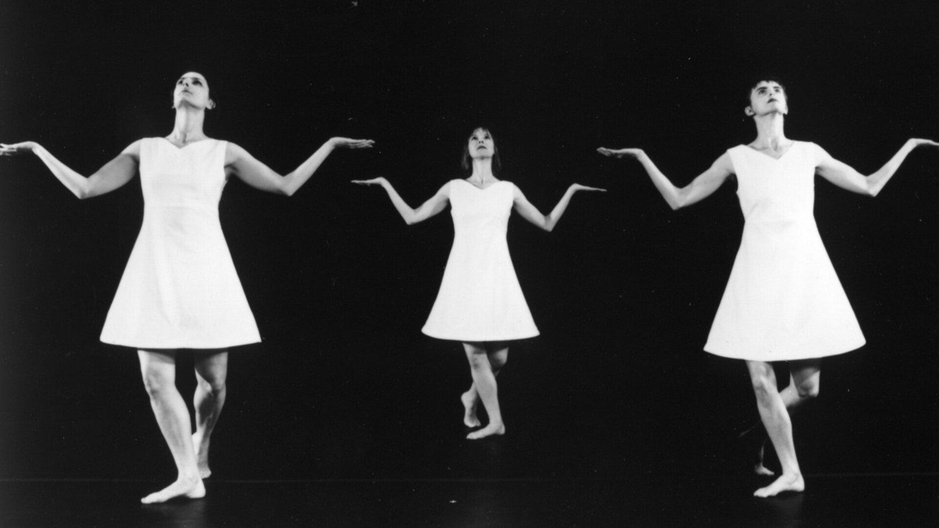 Three dancers in short triangular white dresses stand in identical postures, with arms bend from their sides, palms up and looking up